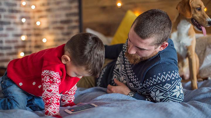 Father, son and pet dog sitting on bed with mobile phone to call Santa's Hotline with Christmas lights in background