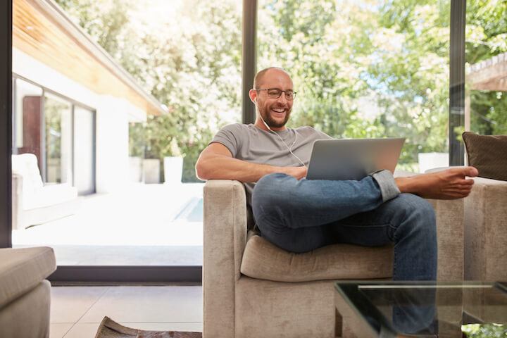 Indoor shot of a happy mature man having video call with laptop and earphones while sitting on the couch at home. Caucasian man using laptop and smiling.